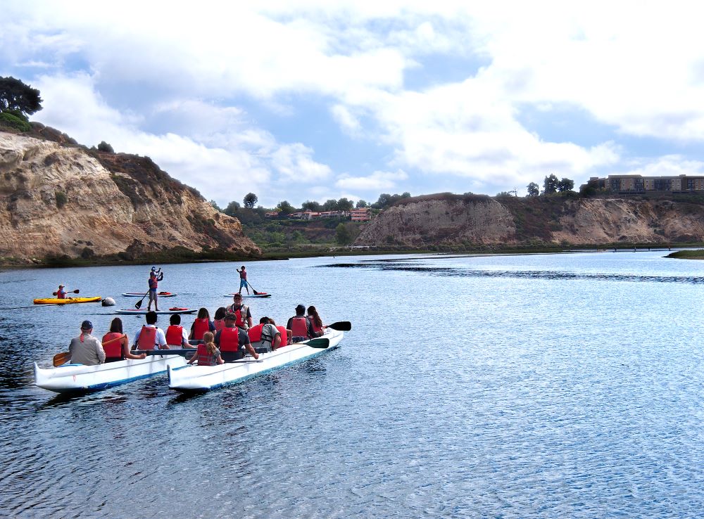 Photo of people in outrigger canoe paddling in Newport's Back Bay, with hills and trees in the background.
