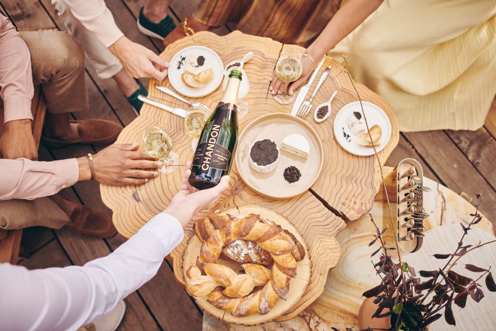 Photo of champaign being poured in glasses on a table with cheese and bread.