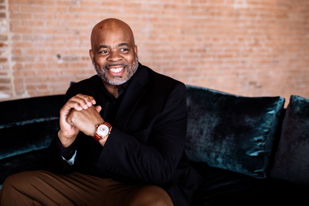 Photo of speaker Andre Norman sitting on a couch in front of a red brick wall.