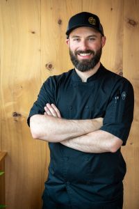 Photo of Chef Michael Chang, standing in a black chef's uniform in front of a wood-paneled wall.