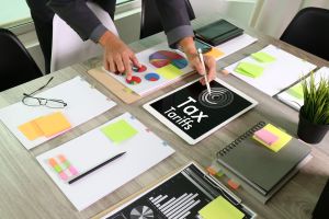 Image of a person with a pencil writing on a piece of paper that reads Tax Tariffs on it, on a desk full of business documents.