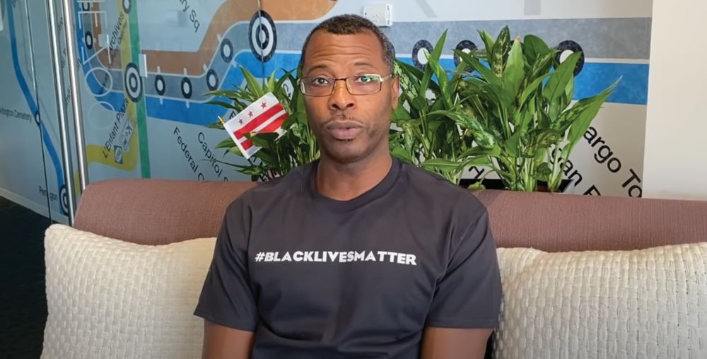 Capture of a still from video with Elliott L. Ferguson II, siting on a couch in a #BlackLivesMatter t-shirt.