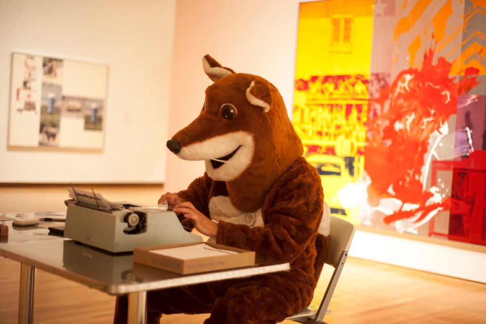 Photo of Poetry Fox, in a fox costume sitting at a typewriter.