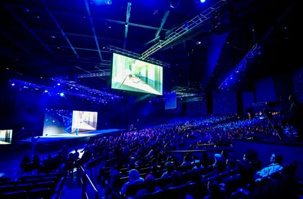 Photo of a group gathered in a large, dark convention hall to see a keynote presentation.