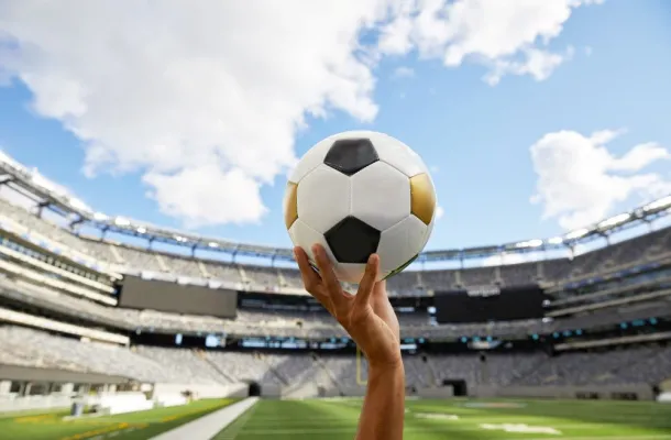 Image of a hand holding a soccer ball with a stadium in the background.