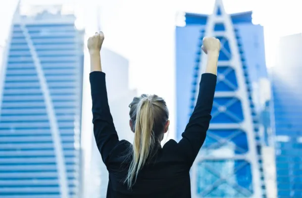 Photo of a woman with her arms up in the air in front of two skyscrapers.