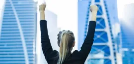 Photo of a woman with her arms up in the air in front of two skyscrapers.