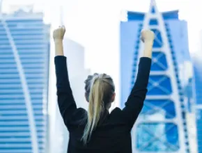 Photo of a woman with her arms up in the air in front of two skyscrapers.