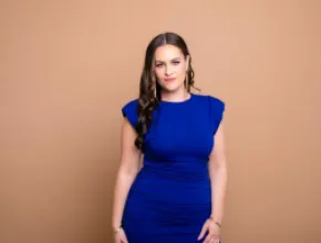 Photo of Courtney Stanley, standing in a blue dress against a beige background.