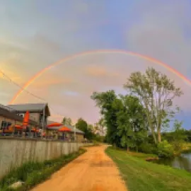 A dirt road along a body of water with a rainbow in the sky.