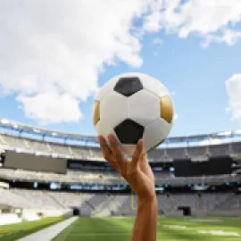 Image of a hand holding a soccer ball with a stadium in the background.