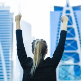 Photo of a woman with her arms up in the air in front of two skyscrapers.