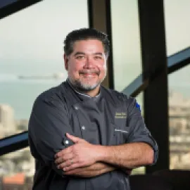 Photo of Chef David Hollands standing in front of a picture window with the skyline of San Francisco in the background.