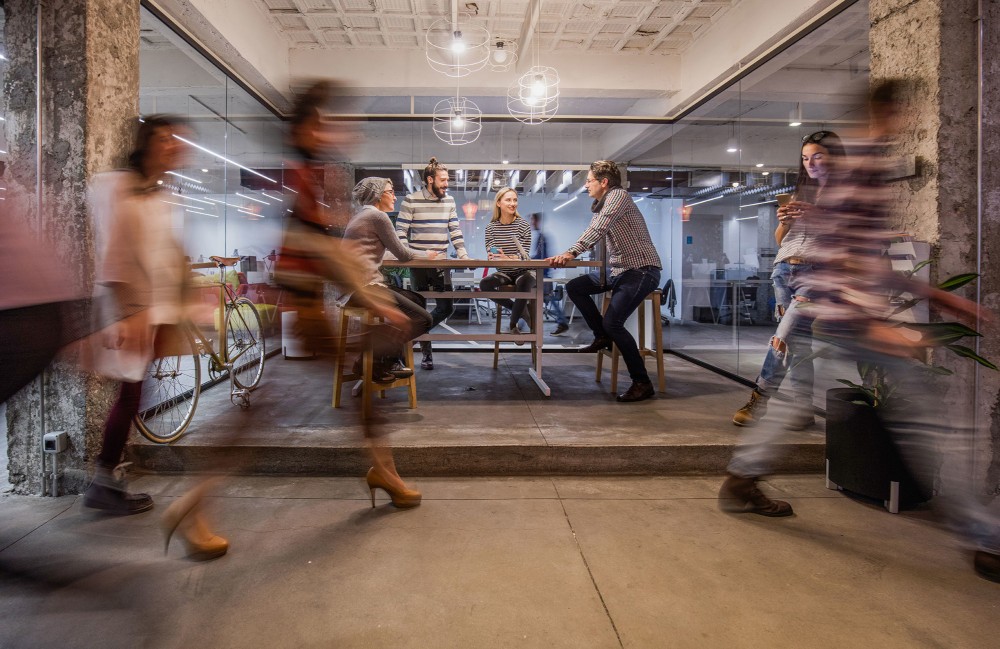 Four people sitting at a table and talking while people walk past.