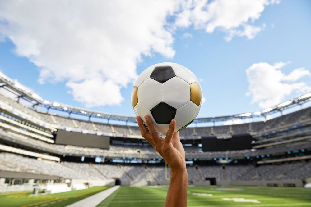 Image of a hand holding a soccer ball with a stadium in the background.