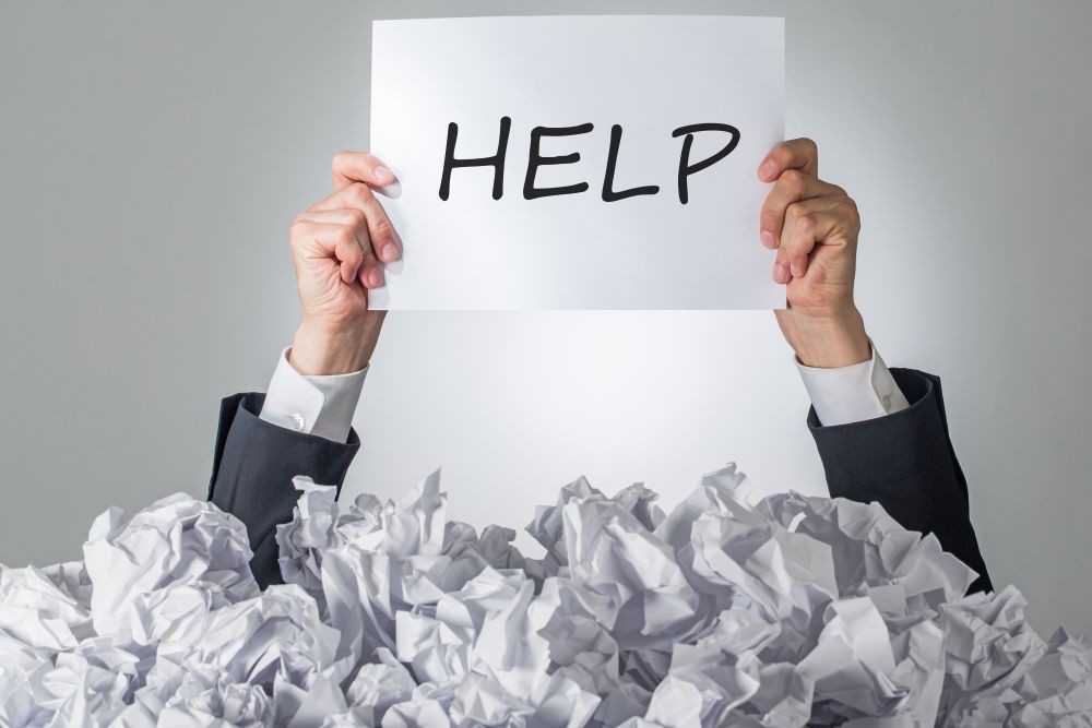 Image of a person holding a paper with the word HELP written on it, emerging from a bunch of crumpled up papers.