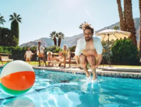 Photo of man jumping into a pool with a beach ball nearby.